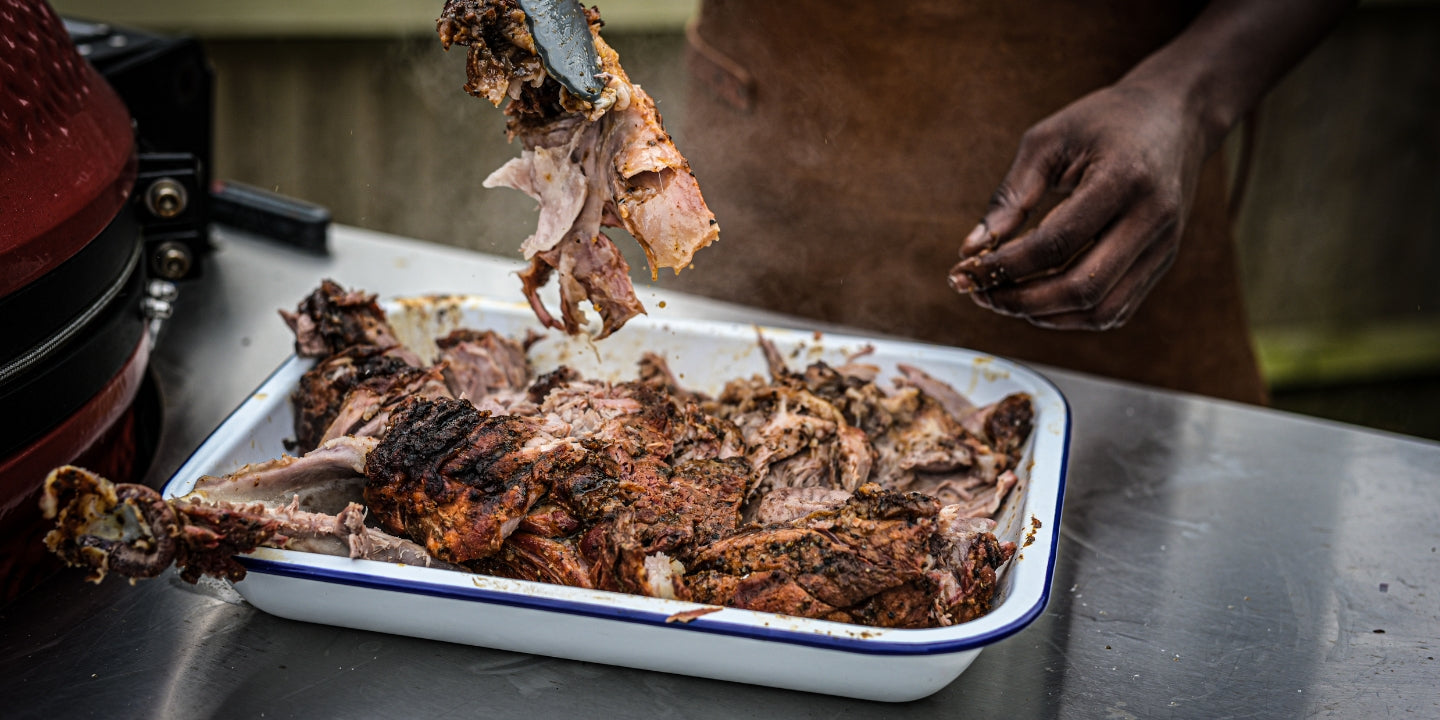 Pulled pork being shredded in an enamel tray after slow cooking on a BBQ, ready to be served in tacos.