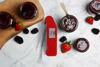 A red Thermapen NANO thermometer with a probe lies on a white marble surface, surrounded by glass jars of berry jam, whole strawberries, and blackberries. One of the jars is open, showing the dark red jam inside, and a spoon rests inside it.