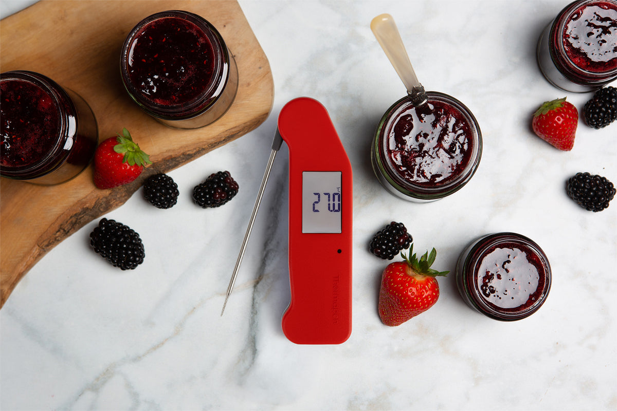 A red Thermapen NANO thermometer with a probe lies on a white marble surface, surrounded by glass jars of berry jam, whole strawberries, and blackberries. One of the jars is open, showing the dark red jam inside, and a spoon rests inside it.
