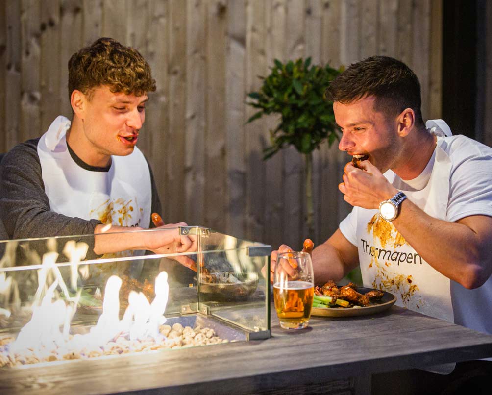 Two men eating a messy barbeque, one wearing a Thermapen apron