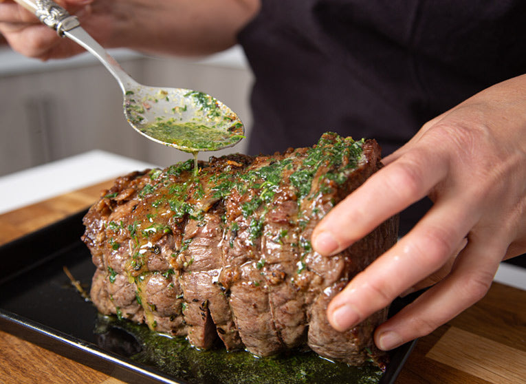 Person seasoning roast topside of beef with tarragon butter on a wooden chopping board.