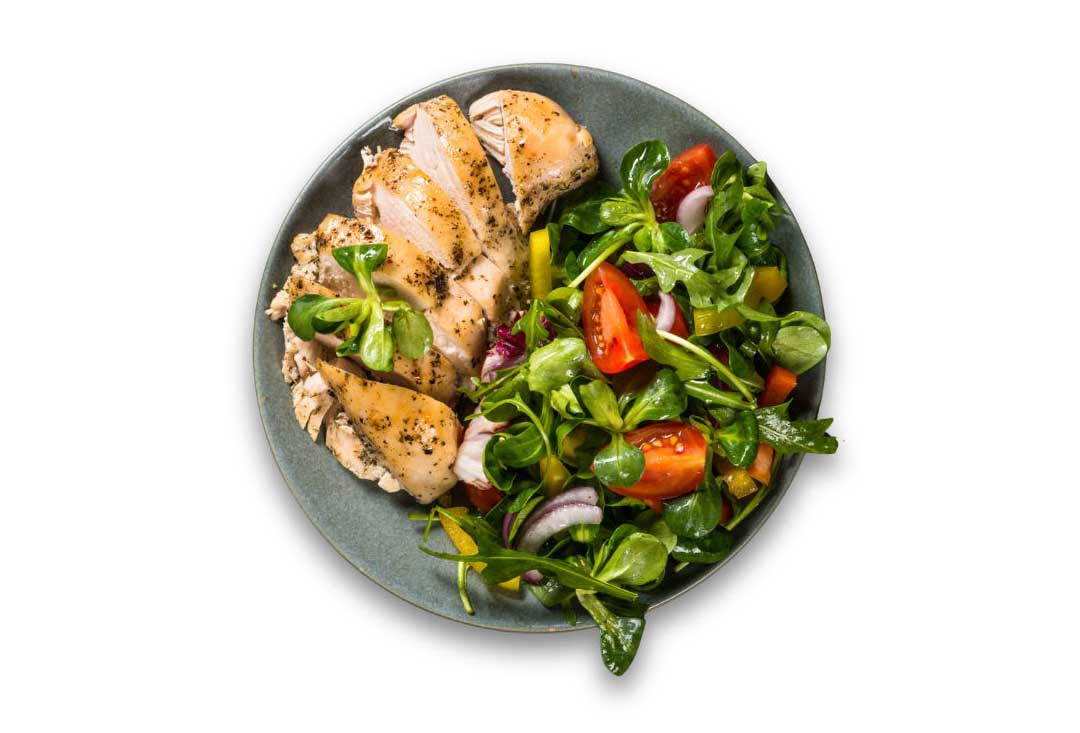A top-down shot of a plate with sliced, roast chicken breast and a fresh green salad, featuring mixed greens, cherry tomatoes, red onions, and yellow bell peppers. Isolated on a white background.