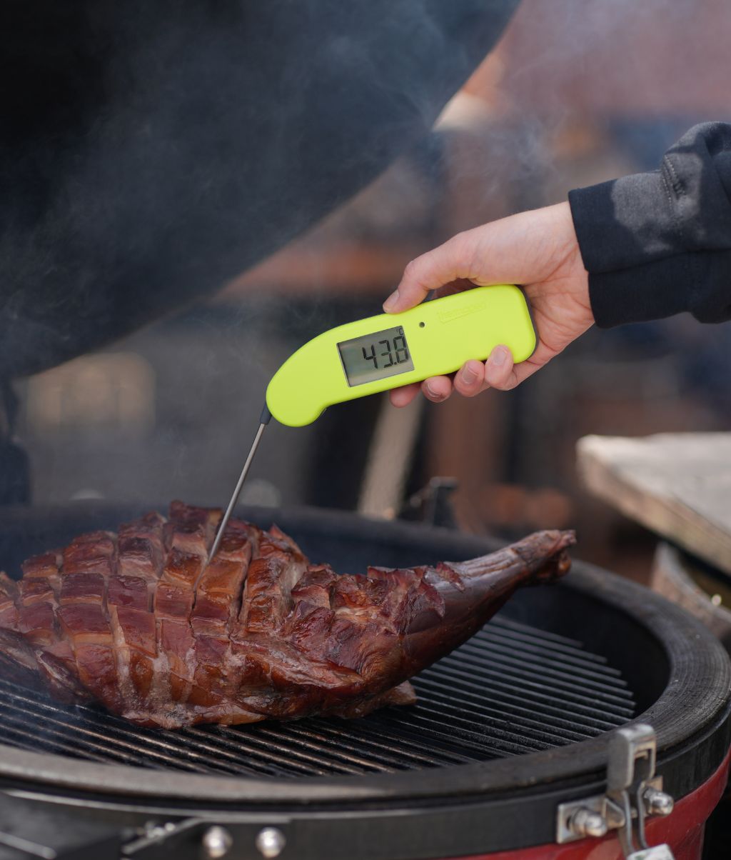 A person measuring the temperature of a lamb leg on a BBQ using a yellow thermapen one thermometer. The temperature display is reading 43.8 degrees 