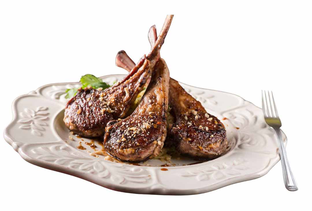 Three perfectly cooked lamb chops, French-trimmed with exposed bones, are elegantly presented on a decorative white plate. The chops have a beautiful seared crust, are sprinkled with what appears to be a nut or breadcrumb topping.