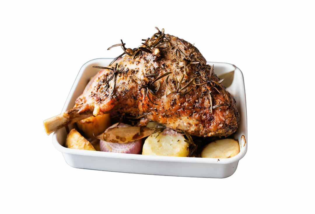 A perfectly roasted leg of lamb, golden brown and crusted with herbs, primarily fresh rosemary sprigs, resting in a white rectangular baking dish. It is surrounded by roasted potatoes and halved red onions, suggesting a delicious Sunday roast.