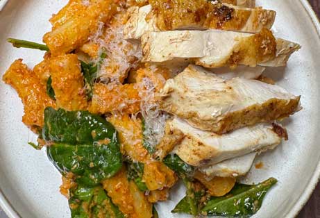 Close-up of marry-me chicken pasta with sliced chicken breast, rigatoni pasta in a creamy orange sauce with spinach, topped with grated Parmesan cheese.