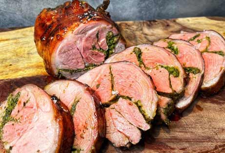 Slices of a roasted, butterflied leg of lamb, revealing a vibrant green herb and garlic stuffing swirled within the tender pink meat. The succulent lamb is arranged on a wooden cutting board with the remaining roast in the background.