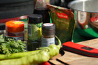 A collection of kitchen ingredients and tools arranged on a wooden cutting board, including a green chili pepper, fresh mint leaves, a bag of Bodrum Pepper Flakes, spice jars, and a red Thermapen NANO thermometer.