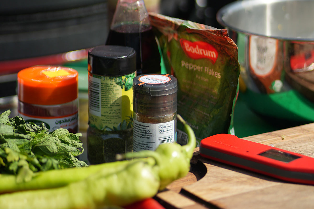 A collection of kitchen ingredients and tools arranged on a wooden cutting board, including a green chili pepper, fresh mint leaves, a bag of Bodrum Pepper Flakes, spice jars, and a red Thermapen NANO thermometer.