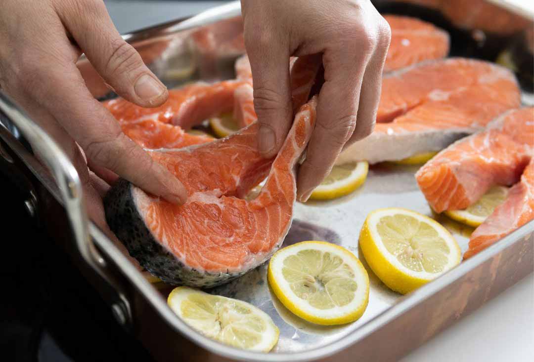 A pair of hands is carefully placing or adjusting a thick cut raw salmon steak with its skin on into a metal baking tray. Several other raw salmon steaks and bright yellow lemon slices are already arranged in the tray, ready for cooking.
