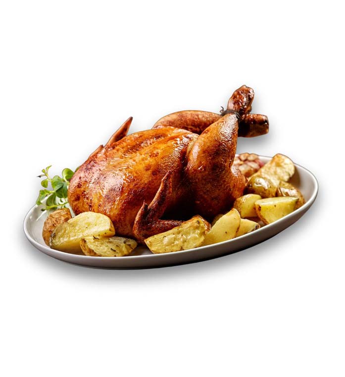 A golden-brown roasted chicken is presented on a white oval platter, surrounded by roasted potato wedges and garlic cloves. And small sprig of fresh herbs as ganish.