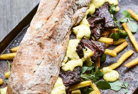 Steak butty with watercress, fries, and creamy mustard sauce on a baguette, served on a metal tray.