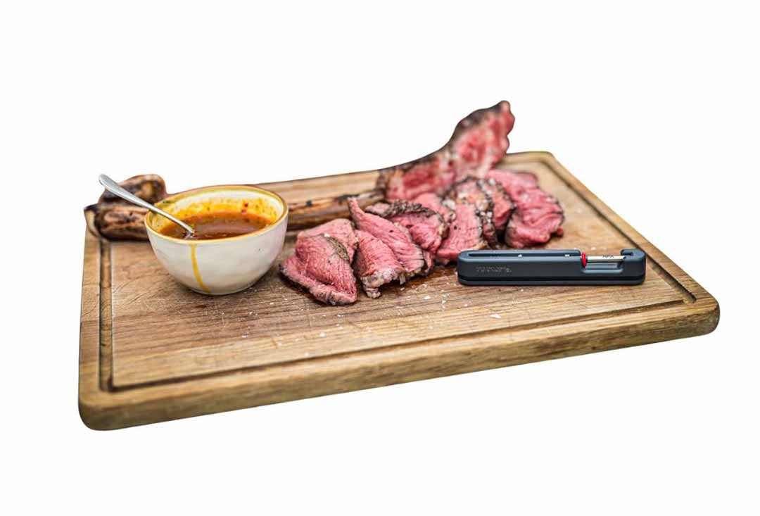 A large tomahawk steak, evenly cooked to a medium-rare doneness with a pink interior, is sliced and fanned out on a rustic wooden cutting board. A small white bowl of orange sauce with a spoon, and a black meat thermometer with a red button, are also on the board.
