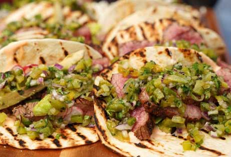 Close-up of grilled steak tacos topped with fresh green salsa and red onions on warm tortillas.