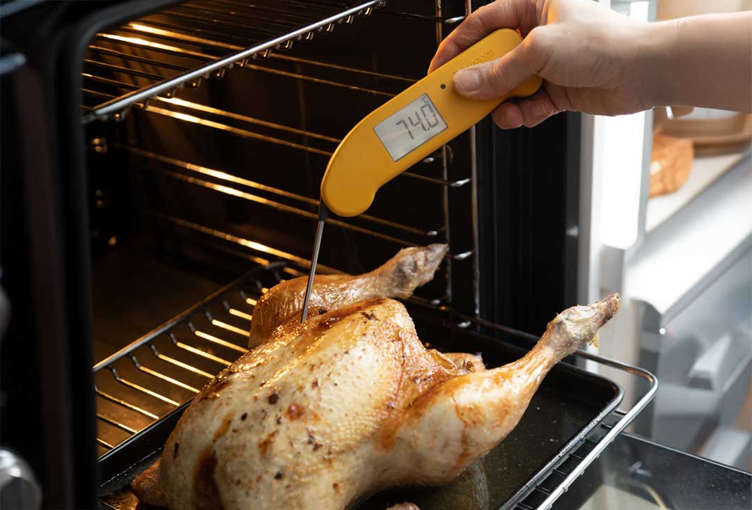 A close-up view shows a hand holding a yellow Thermapen ONE digital thermometer, with a reading of "74.0" degrees Celsius, inserted into the breast of a golden-brown roasted chicken inside a hot oven.