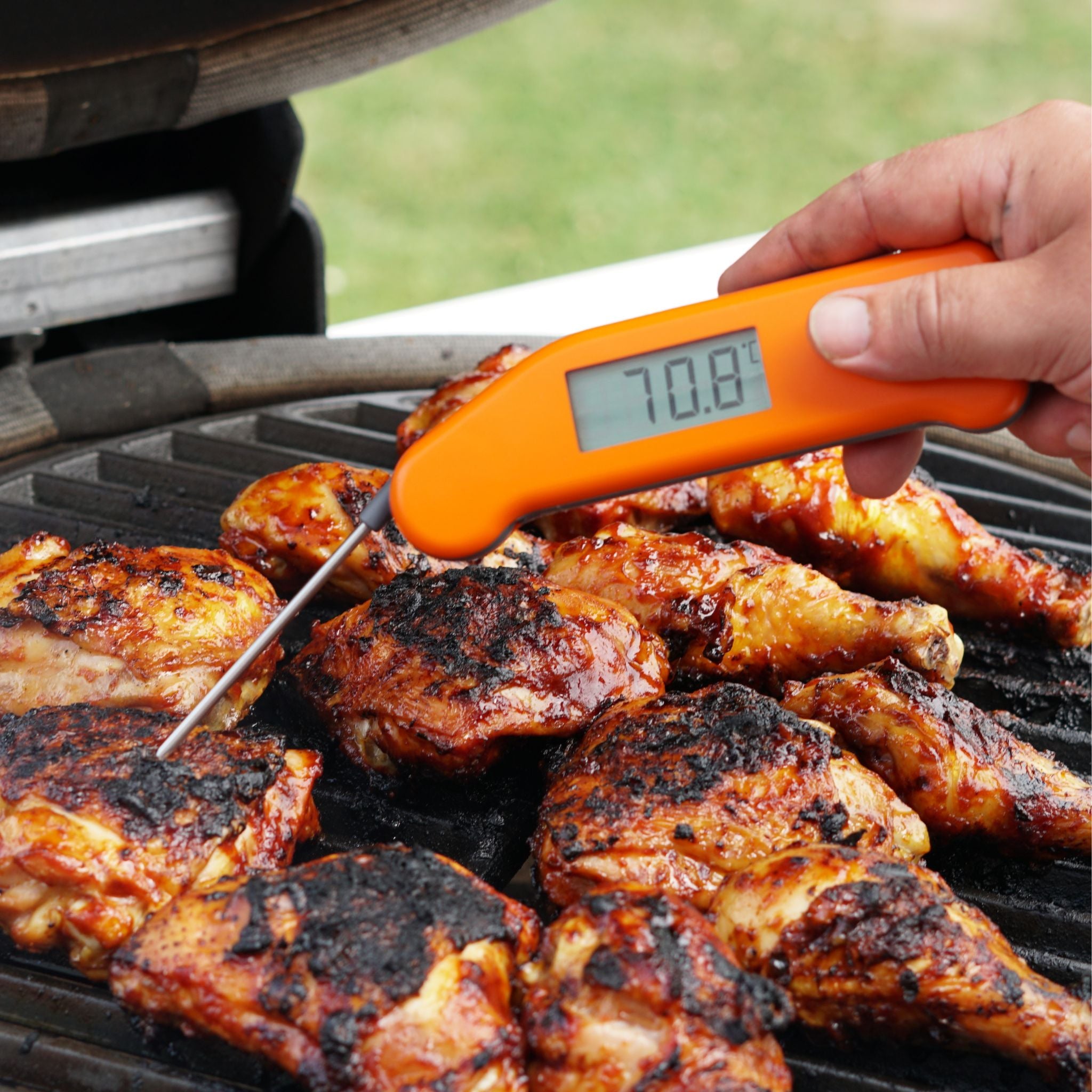 Person using an orange digital thermometer to check the temperature of grilled chicken on a barbecue.