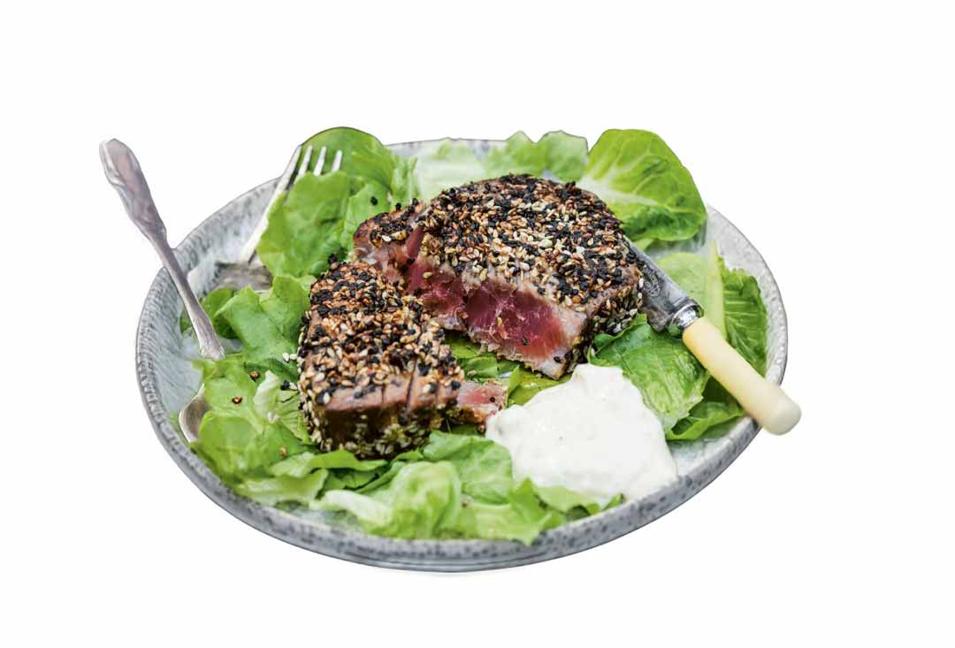 Two thick slices of seared tuna steak, coated generously with a black and white sesame seed crust, showcasing a vibrant rare red interior. The tuna is served on a bed of fresh green lettuce leaves with a dollop of white sauce (possibly aioli or yogurt) and is accompanied by a fork and a knife with an ivory handle, all on a light grey plate.