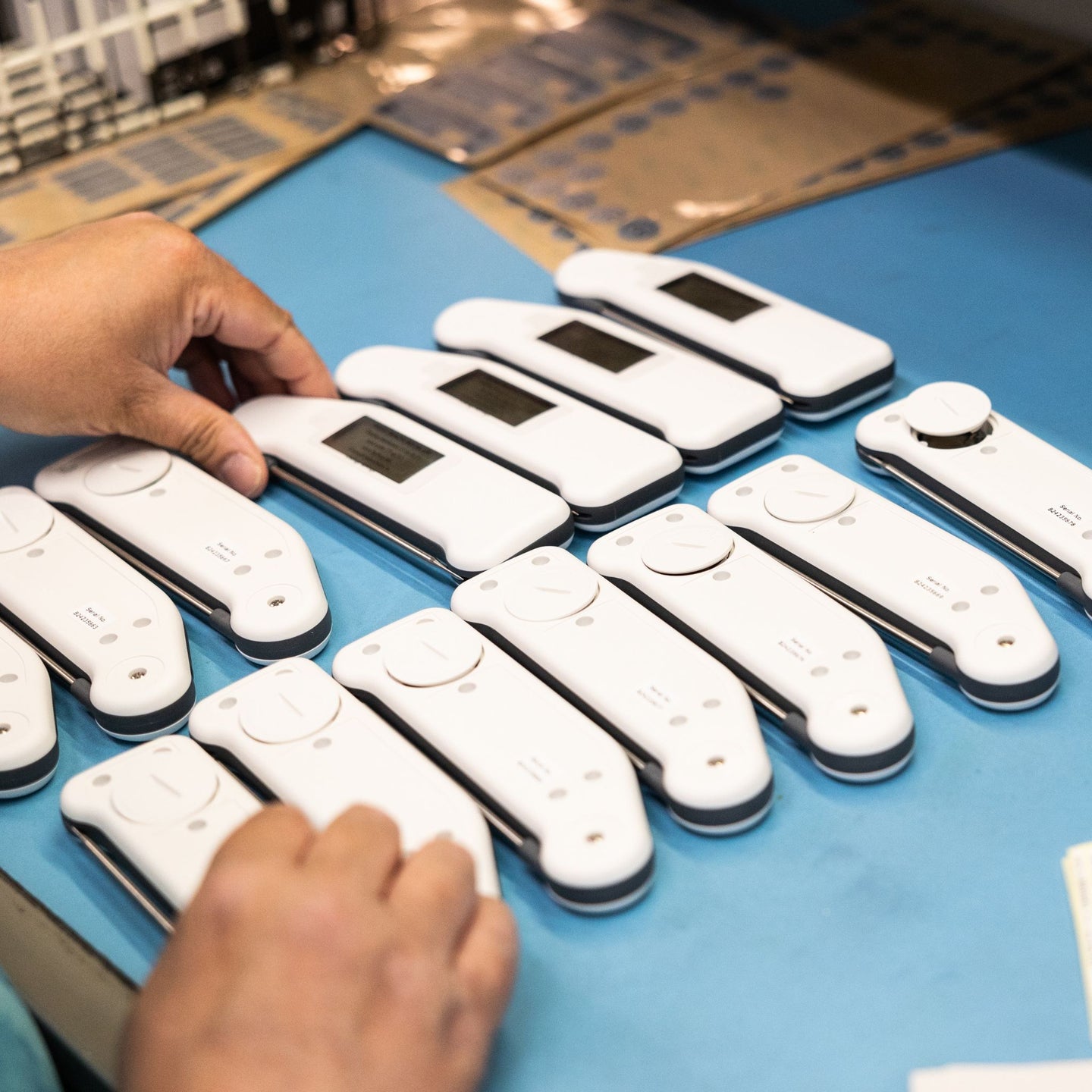 Multiple white thermometers on a blue table, with hands mid build process.