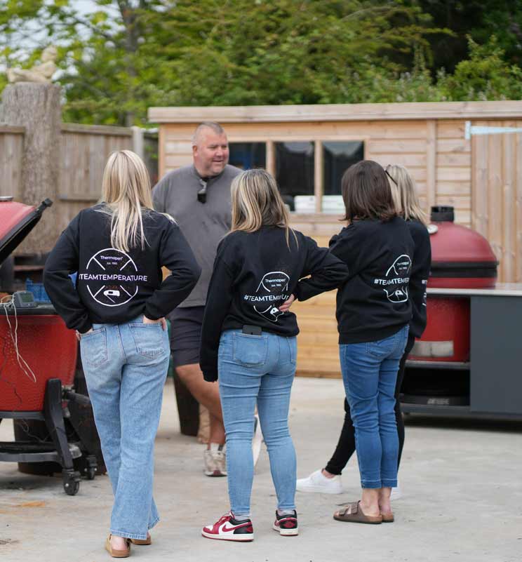 the smokin elk's bbq class in action with 4 women wearing team temperature jumpers gathered around to learn from elky at his bbq shack with kamado joe's in the background