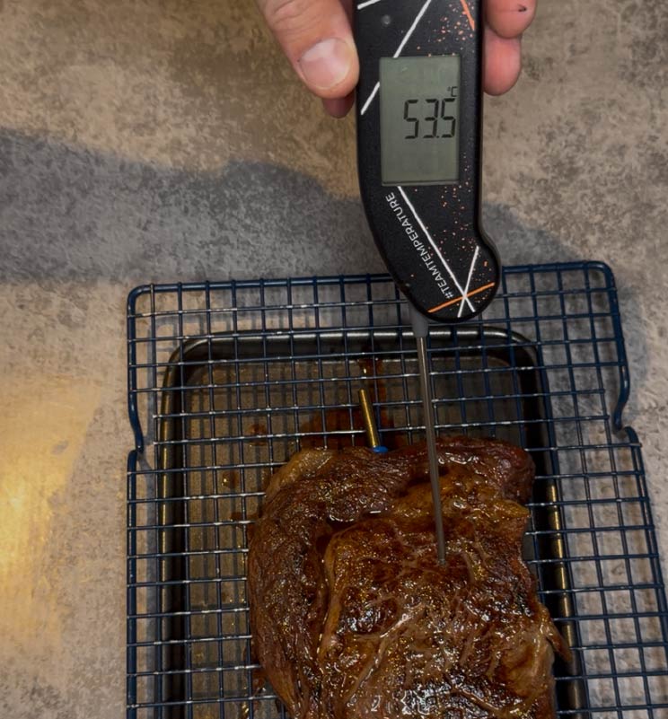 Close-up of a Team Temperature design Thermapen thermometer precisely measuring the internal temperature of a cooked ribeye steak resting on a wire rack, displaying 53.5°C (128.3°F).