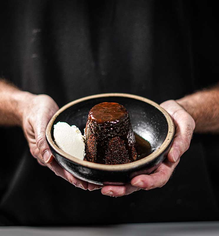 Hands holding plated sticky toffee pudding with cream on the side and sauce poured over the top