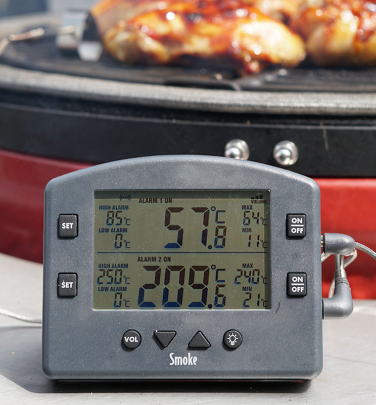 A Smoke thermometer in front of pieces of chicken on a BBQ