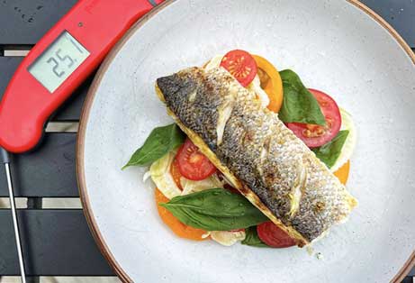 A cooked sea bass fillet with crispy skin, served on a plate with sliced tomatoes, basil leaves, next to a red Thermapen digital meat thermometer 