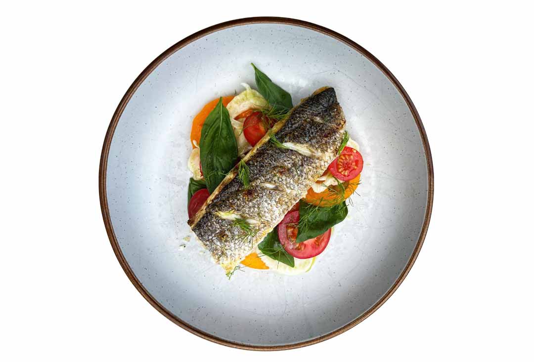 An overhead shot of a crispy-skinned whole cooked fish fillet, sea bass, served on a light grey plate with a rustic brown rim. The fish is garnished with fresh dill and rests atop a colorful salad of bright orange slices, vibrant green basil leaves, and sliced red tomatoes.