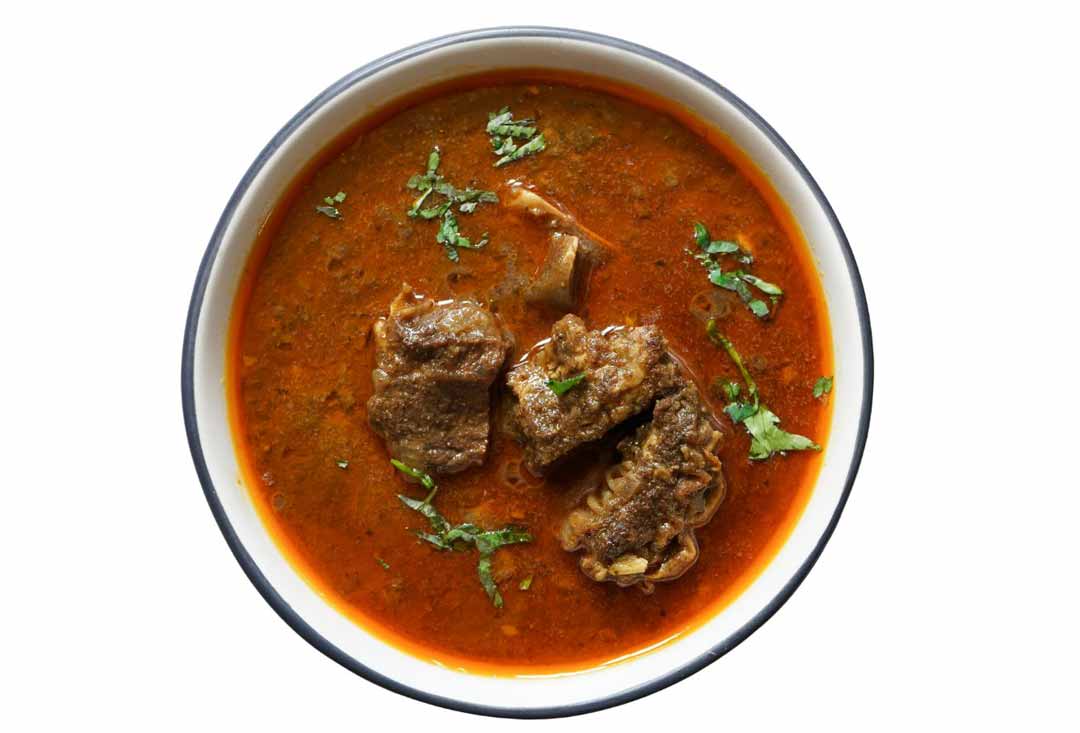 A golden-brown roasted chicken is presented on a white oval platter, surrounded by roasted potato wedges and An overhead shot of a rich, reddish-brown lamb curry or stew served in a white bowl with a dark rim. Several tender pieces of lamb meat, some still on the bone, are visible in the thick gravy, garnished with fresh chopped coriander leaves. cloves. And small sprig of fresh herbs as ganish.