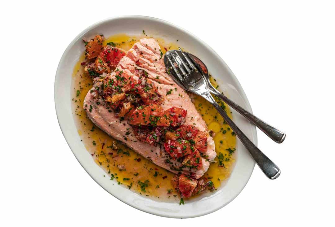 An overhead view of a perfectly cooked salmon fillet, light pink in color, served on an oval white plate. It's topped with a vibrant red and orange salsa made from what appears to be citrus segments and herbs, all resting in a golden butter or olive oil sauce. A fork and spoon are laid neatly beside the salmon.