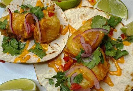 Two fish tacos made with crispy battered fish, topped with red onion slices, cilantro, and a creamy orange sauce, served on tortillas with lime wedges on the side.