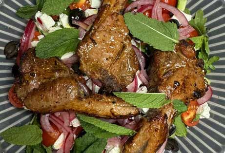 Plate of grilled lamb chops, lightly seasoned with what appears to be sumac, served over a vibrant salad of red onion, tomatoes, black olives, and crumbled feta cheese, garnished with fresh mint leaves.