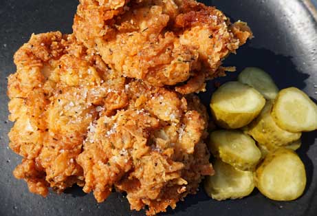 Overhead view of two pieces of golden-brown, crispy fried chicken, sprinkled with seasoning, next to a pile of sliced dill pickles on a dark plate.