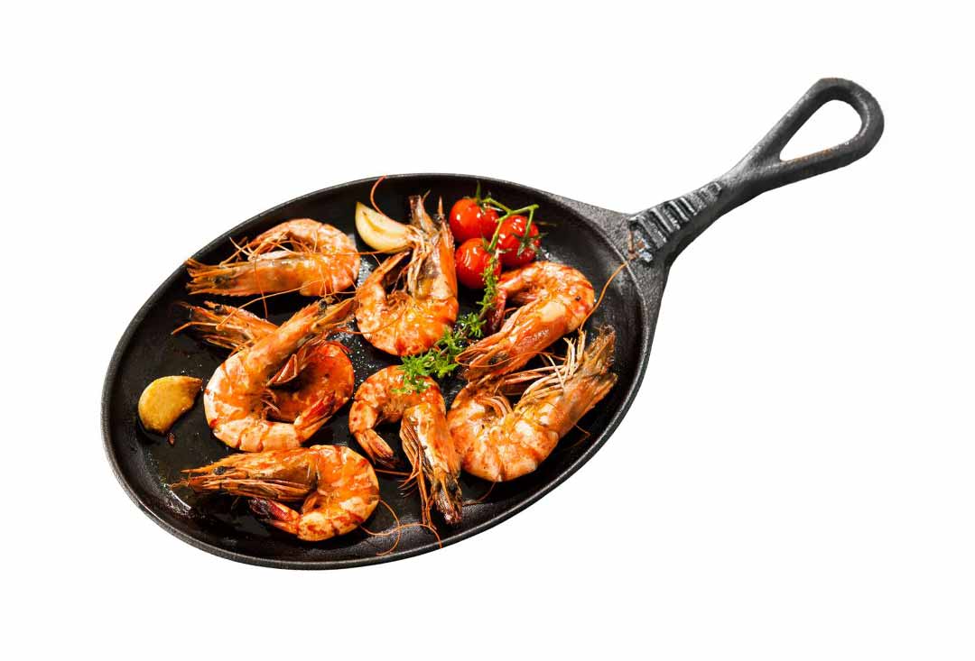 An overhead shot of several grilled whole king prawns with their shells and heads on, showing an appealing orange and pink color, served in a black cast iron skillet. The prawns are accompanied by roasted garlic cloves, a small cluster of red cherry tomatoes on the vine, and fresh green herbs.