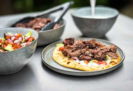 Close-up of shredded lamb served on a flatbread with a creamy white sauce and diced tomato and cucumber salad. In the background, a bowl of more shredded lamb and a bowl of the white sauce are out of focus on a metal table, suggesting an outdoor meal.