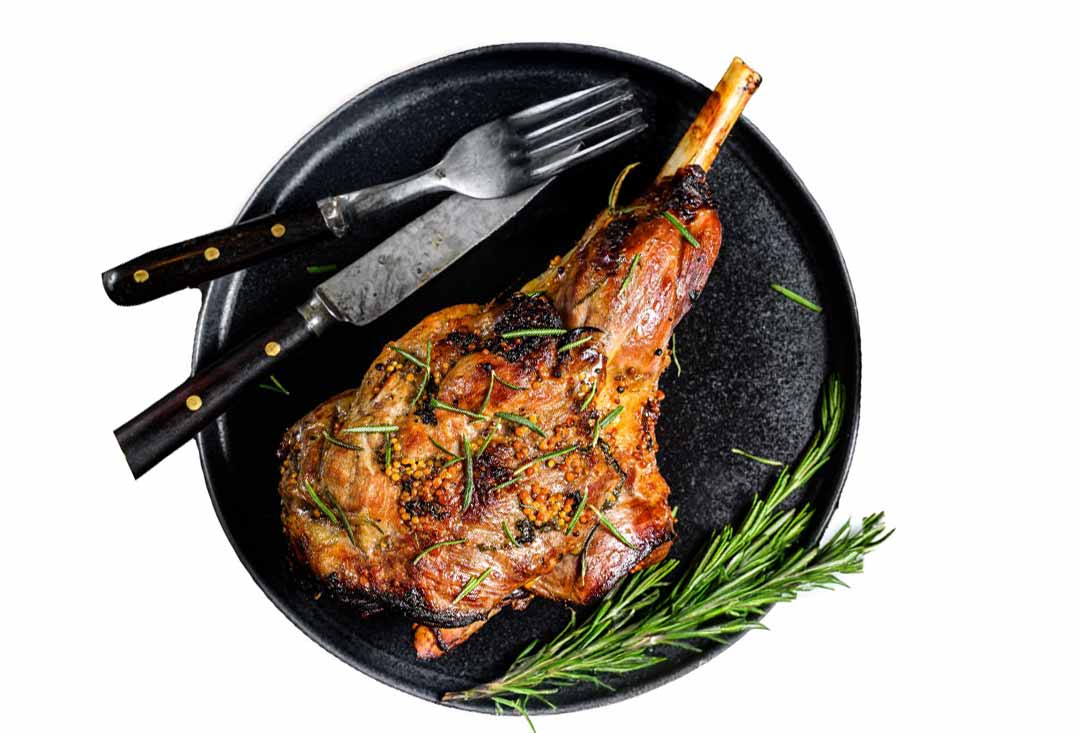 An overhead view of a beautifully roasted leg of lamb, golden brown with a crispy skin, generously seasoned with fresh rosemary sprigs and mustard seeds. It's presented on a matte black plate alongside a vintage-style fork and knife with dark handles, and an extra sprig of rosemary.