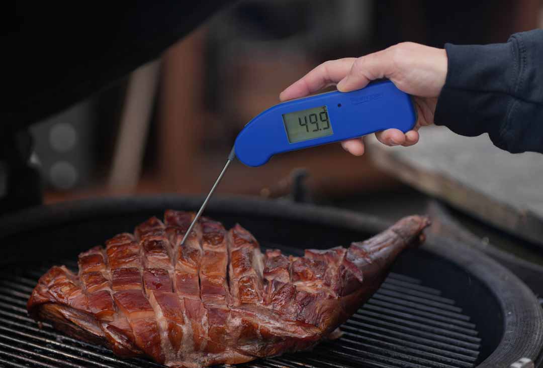 A hand holds a blue digital meat thermometer, displaying '49.9' degrees Celsius, with its probe inserted into a large, scored leg of lamb cooking on a barbecue grill. The lamb has a rich, deep brown crust from the grilling process.