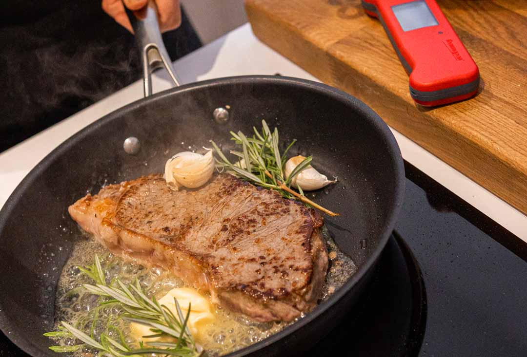 An overhead shot of a seared steak cooking on a black skillet, surrounded by garlic cloves, fresh sprigs of rosemary all simmering in juices and oil with a red Thermapen by the side