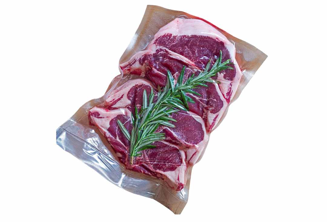 Two raw, red meat steaks, possibly lamb chops or small steaks, vacuum-sealed in a clear plastic bag, with a fresh sprig of rosemary placed on top of the meat, ready for sous vide cooking. The white fat marbling is visible along the edges of the meat.