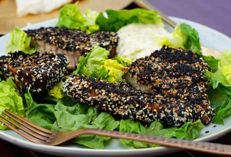 Three seared tuna steaks heavily crusted with black and white sesame seeds, served on a bed of green lettuce leaves.