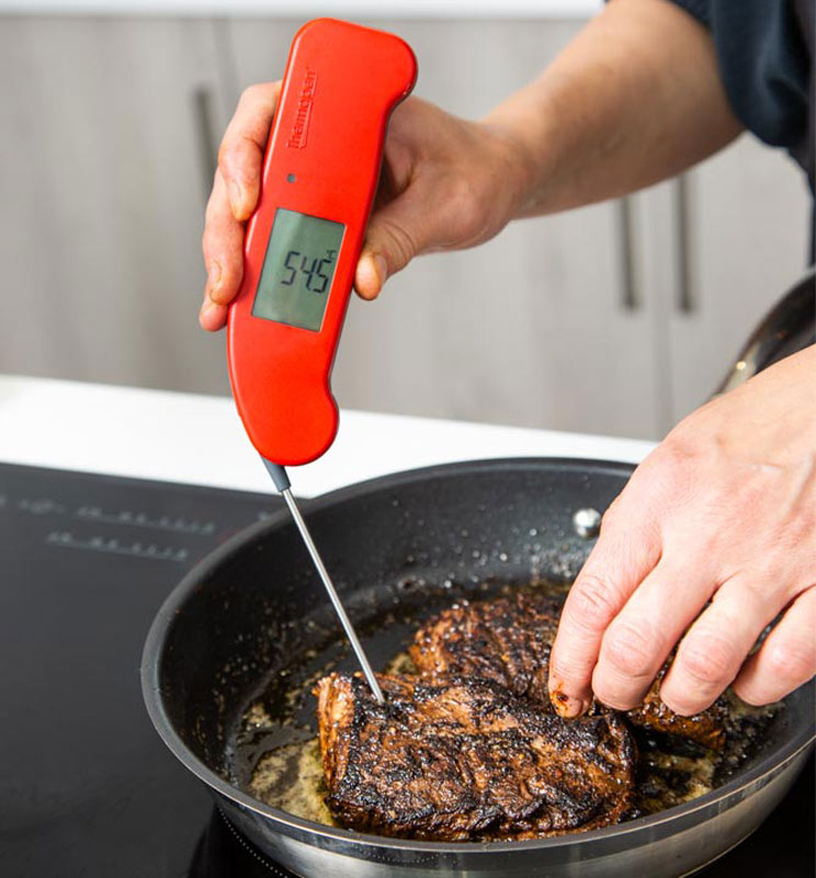 Person measuring steak in a pan with a red Thermapen thermometer