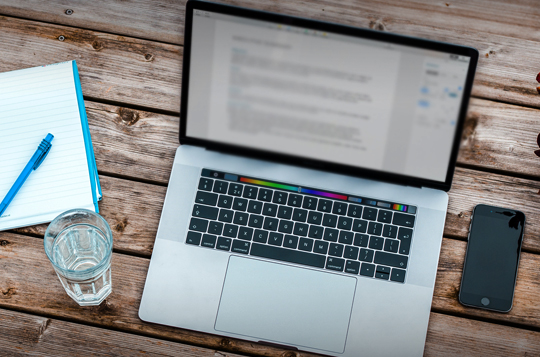 A Birdseye view of a mac on a wooden table with an iPhone and glass of water either side