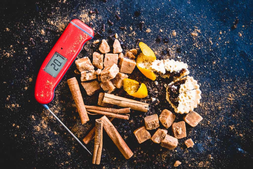 A red Thermapen digital thermometer laid out on a black board with cinnamon sticks and fudge