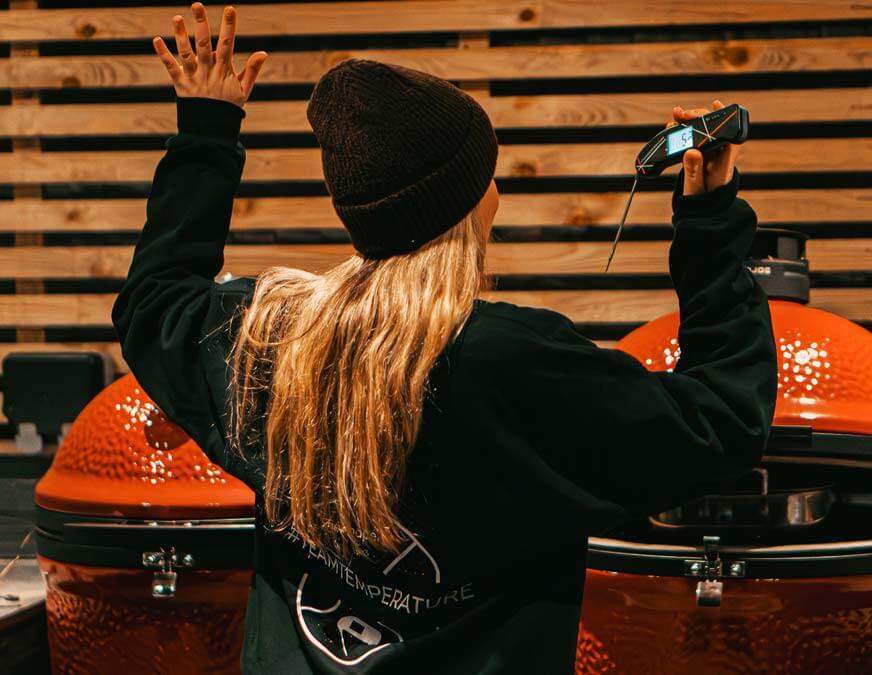 A girl holding Team Temperature Thermapen up in an outdoor BBQ shack wearing Team Temperature sweater and a hat