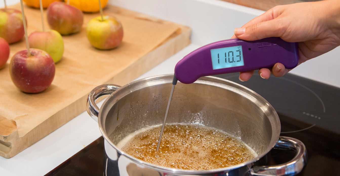 A purple Thermapen ONE is used to measure the temperature of a bubbling pot of caramel or toffee on a stovetop. The thermometer's digital display shows a temperature of 110.3°C. In the background, there are several caramel-covered apples on sticks.