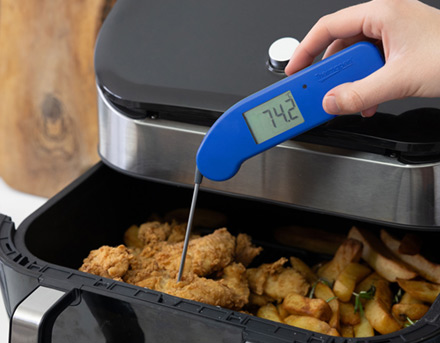 A hand holding a limited edition blue Thermapen ONE instant-read thermometer, measuring the internal temperature of crispy chicken in an air fryer basket, next to golden chips. The display shows 74.2 degrees.